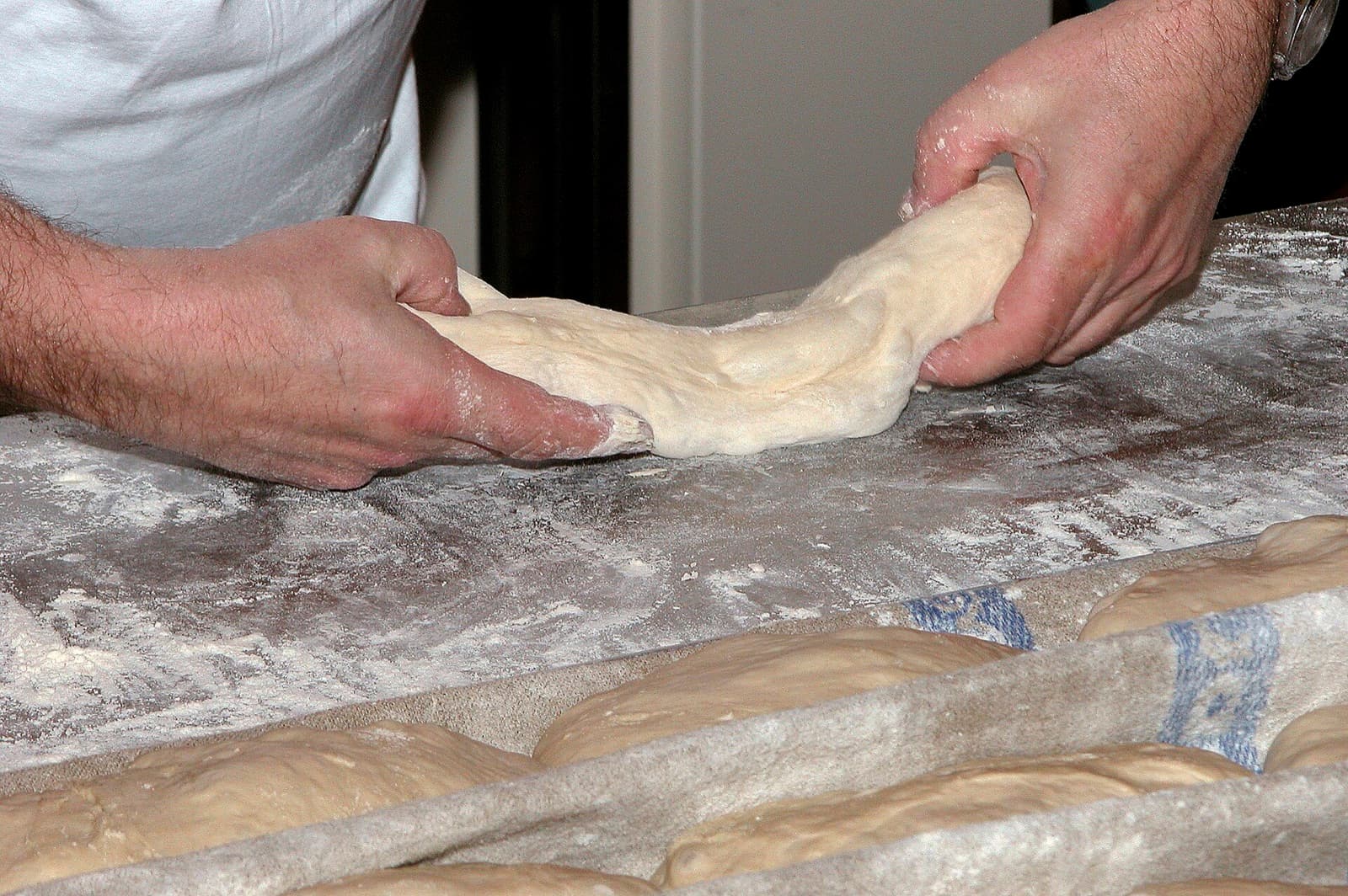 Un boulanger façonne une baguette sur un plan de travail fariné, devant des pâtons sur couche